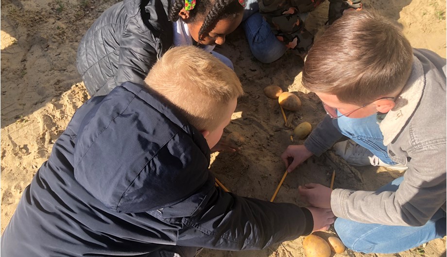 Hunebedden namaken buiten in de zandbak met een zak aardappelen als alternatief voor de stenen. Een mooi voorbeeld van ons middagonderwijs.